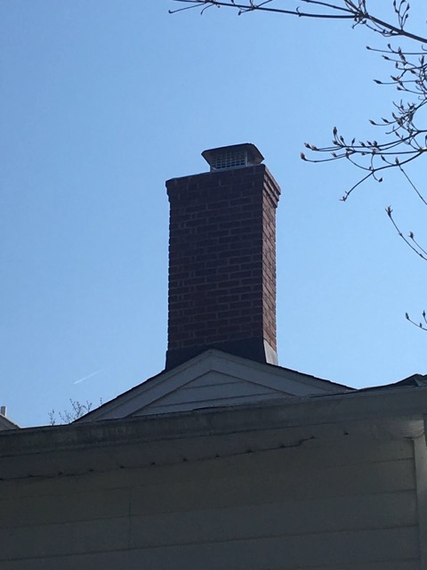 Tall brick chimney with a new metal cap rising above a rooftop against a clear blue sky