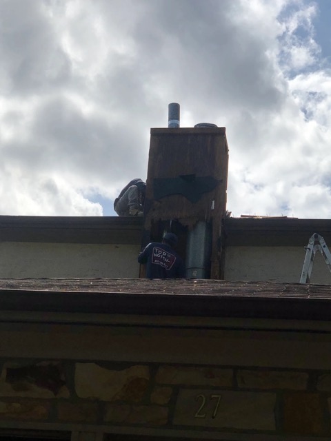 A worker on a rooftop repairing a chimney with an exposed metal flue pipe, ladder resting at the roof edge