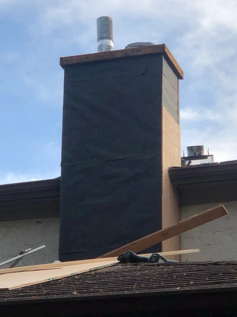 Chimney wrapped in black tar paper with a metal flue pipe rising from it, mid-construction on a shingled roof