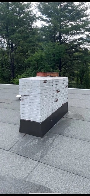 White-painted brick chimney with clay flue tiles and black base flashing surrounded by tall green trees